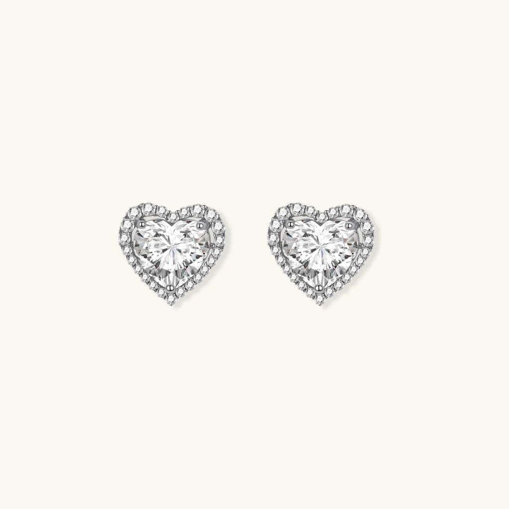 Close-up of heart-shaped moissanite stud earrings in sterling silver halo setting on cream background.