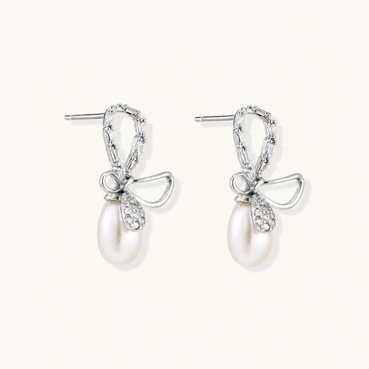 Silver bow-shaped stud earrings with baguette crystals and dangling white freshwater pearls on cream background