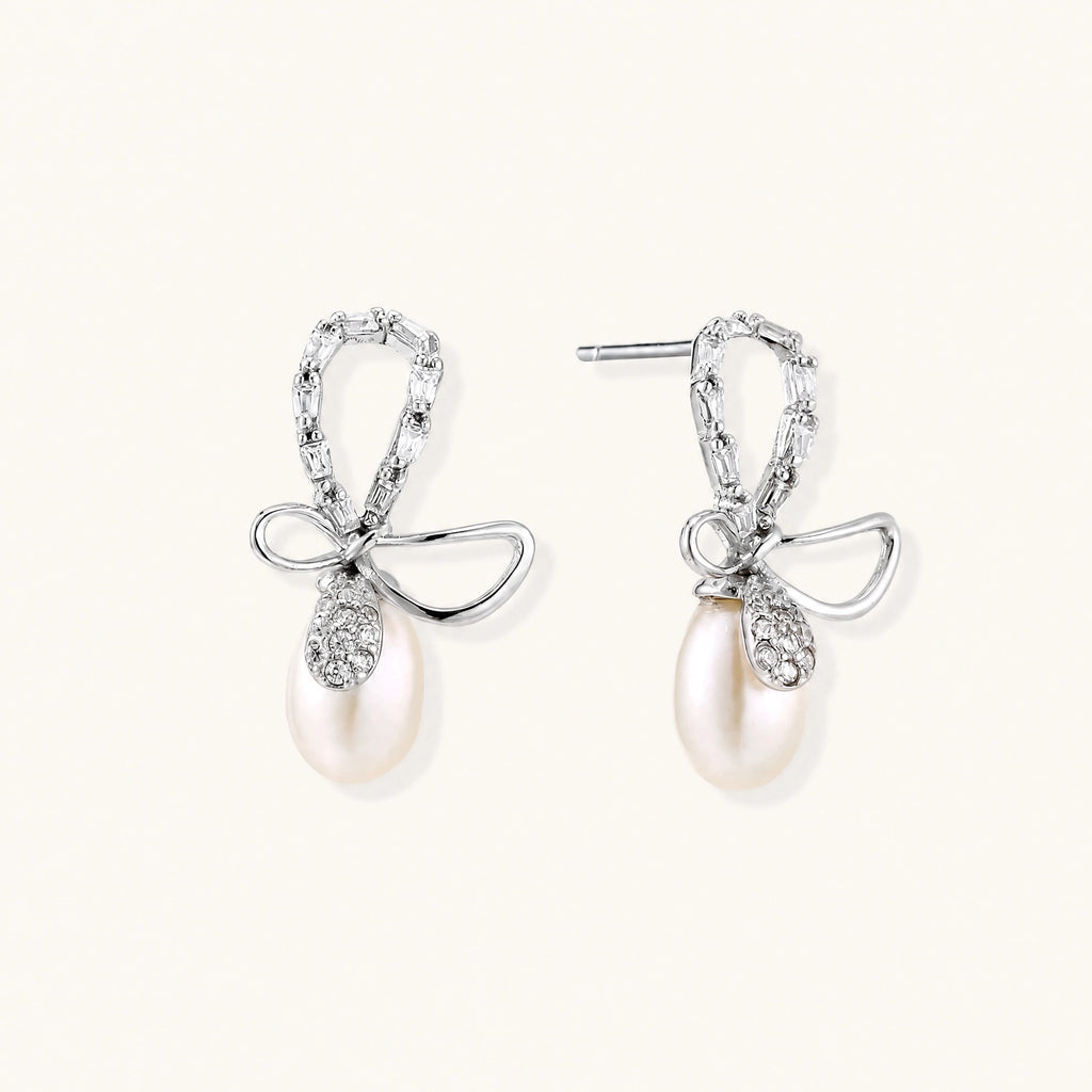Silver bow-shaped stud earrings with baguette crystals and dangling white freshwater pearls on cream background