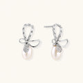 Silver bow-shaped stud earrings with baguette crystals and dangling white freshwater pearls on cream background