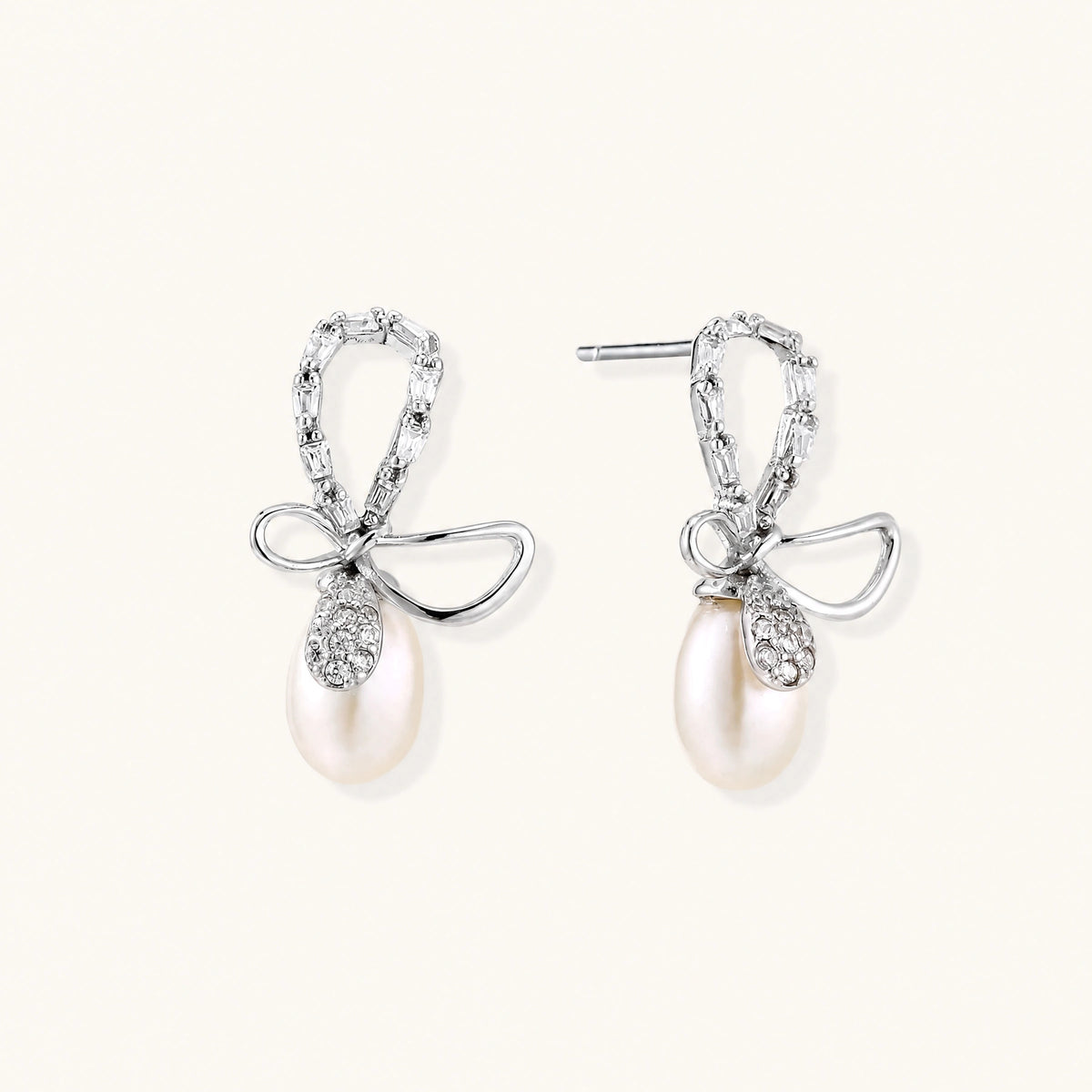 Silver bow-shaped stud earrings with baguette crystals and dangling white freshwater pearls on cream background