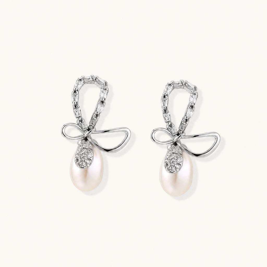 Silver bow-shaped stud earrings with baguette crystals and dangling white freshwater pearls on cream background