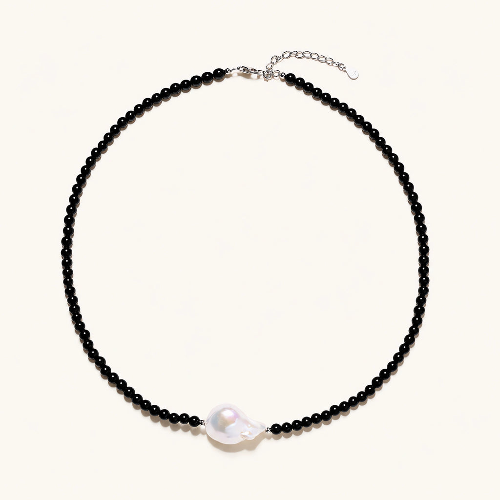 Beaded black agate necklace with iridescent baroque freshwater pearl centerpiece on cream background.