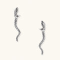 Silver-toned serpent-shaped dangle earrings with detailed scale texture curving in S-shape on beige background