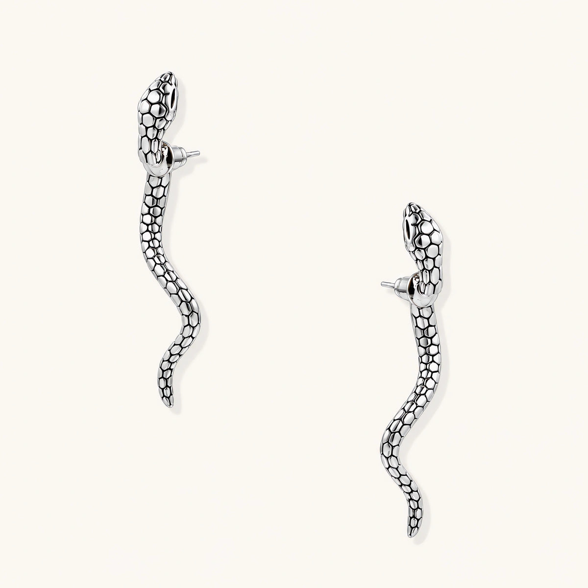 Silver-toned serpent-shaped dangle earrings with detailed scale texture curving in S-shape on beige background