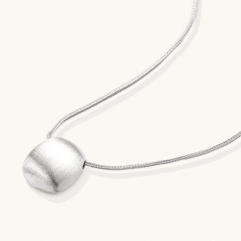 Minimalist brushed sterling silver dome pendant on delicate chain against cream background, showing matte textured finish
