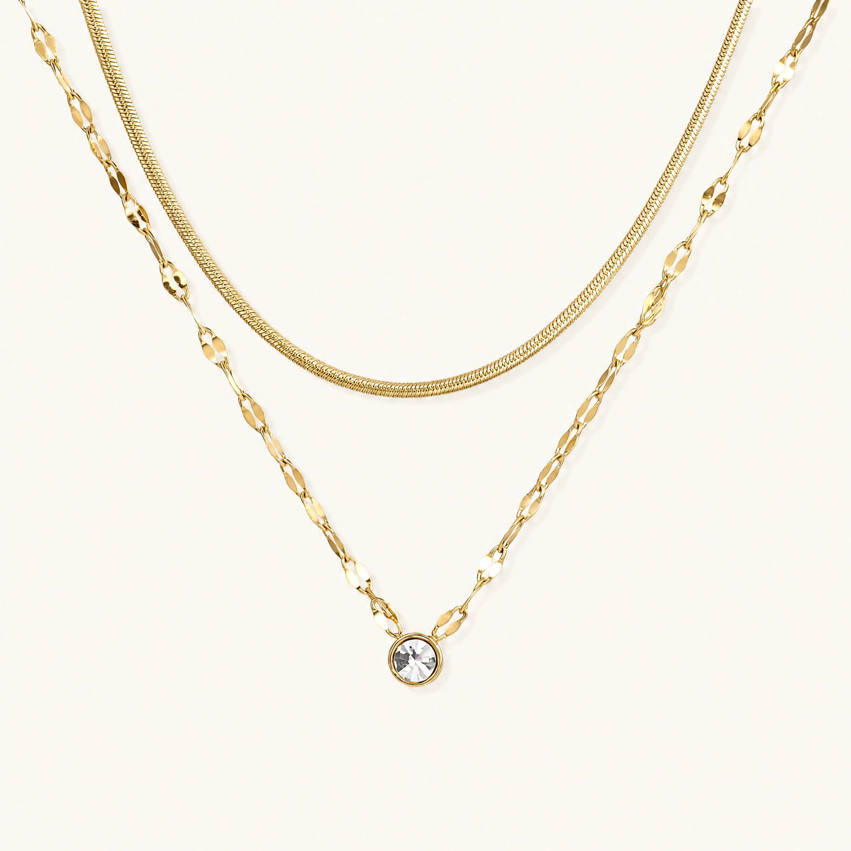 Layered gold necklace with herringbone and link chains, small bezel-set crystal pendant on cream background