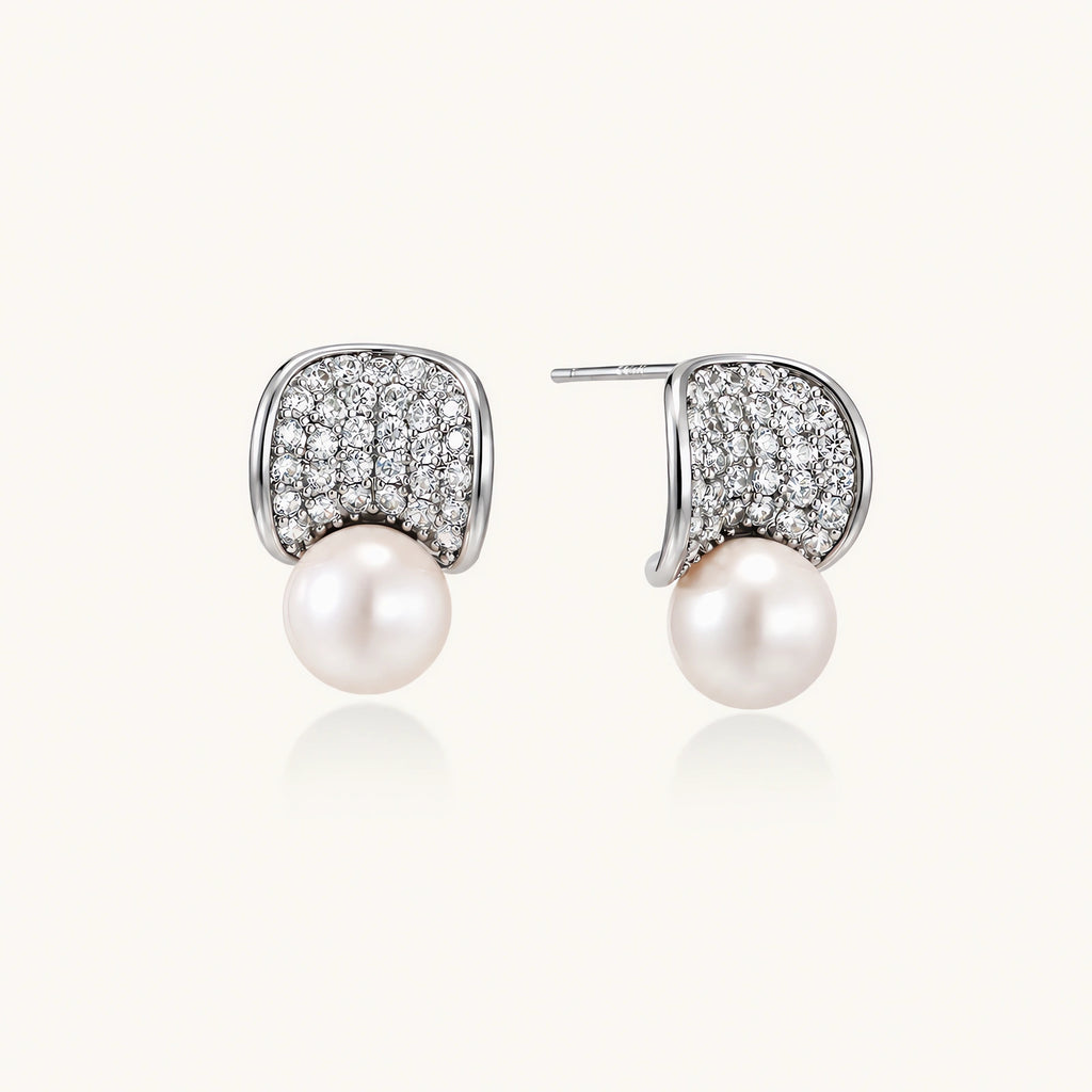 Pair of white gold plated eco-brass stud earrings with curved sparkling stone panels and round freshwater pearls on a cream background.