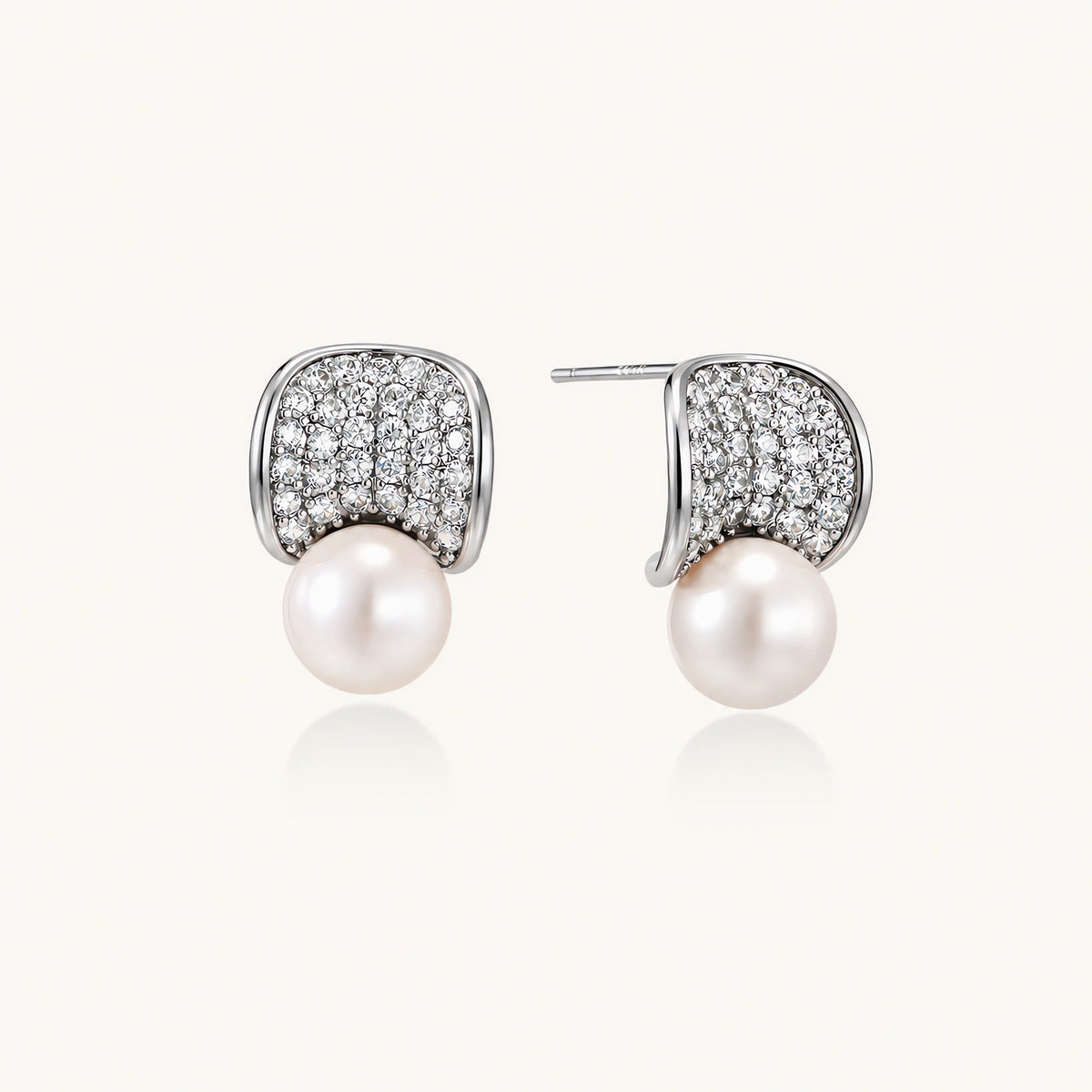 Pair of white gold plated eco-brass stud earrings with curved sparkling stone panels and round freshwater pearls on a cream background.