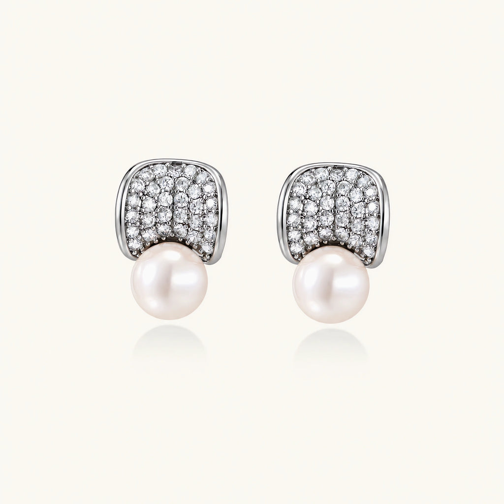 Pair of white gold plated eco-brass stud earrings with curved sparkling stone panels and round freshwater pearls on a cream background.