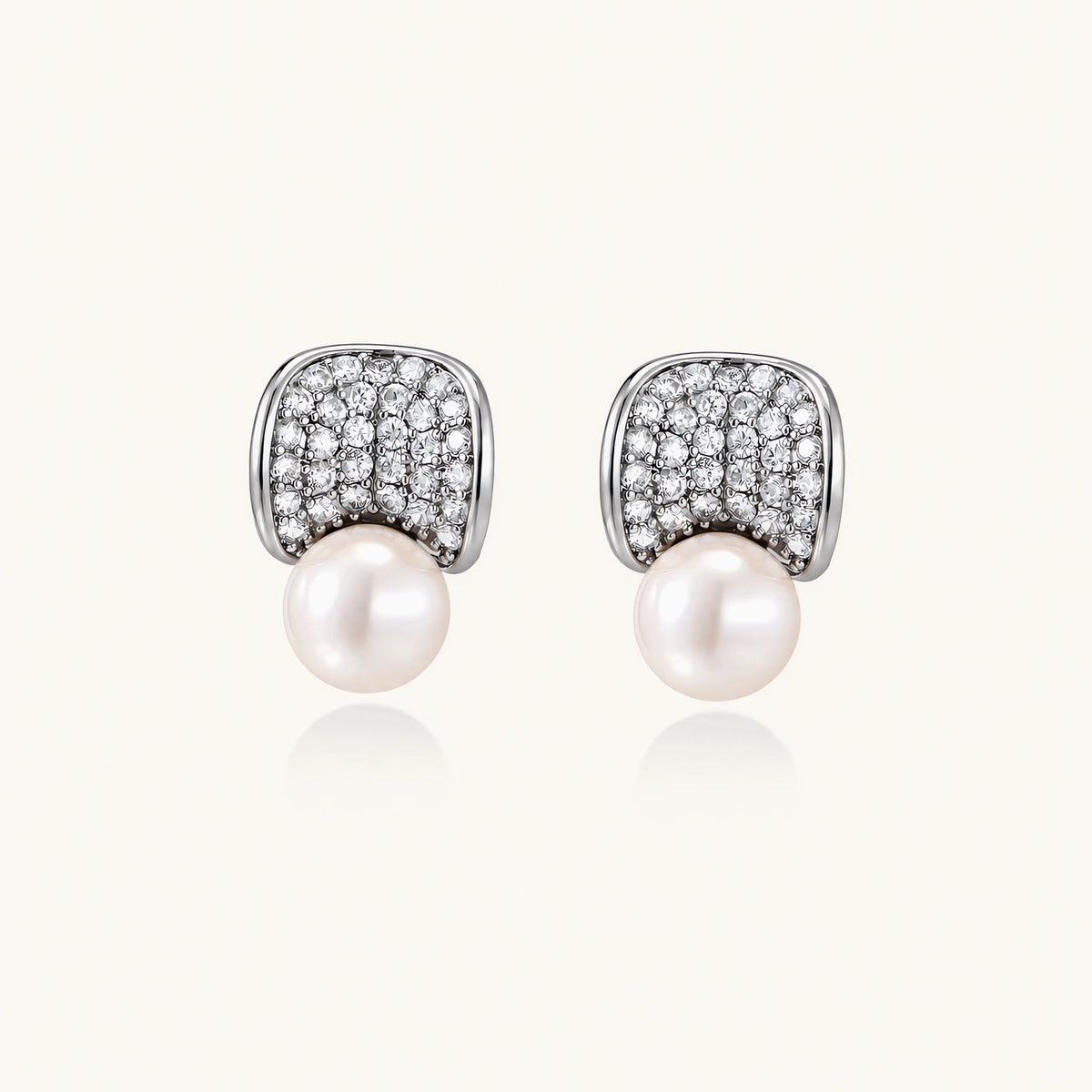 Pair of white gold plated eco-brass stud earrings with curved sparkling stone panels and round freshwater pearls on a cream background.