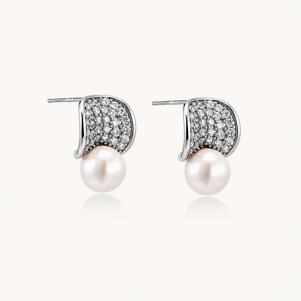 Pair of white gold plated eco-brass stud earrings with curved sparkling stone panels and round freshwater pearls on a cream background.