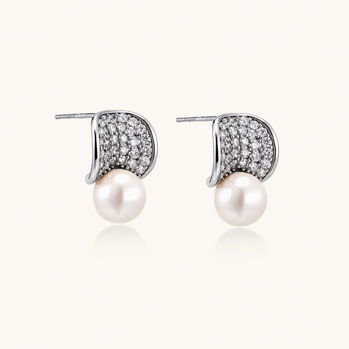 Pair of white gold plated eco-brass stud earrings with curved sparkling stone panels and round freshwater pearls on a cream background.