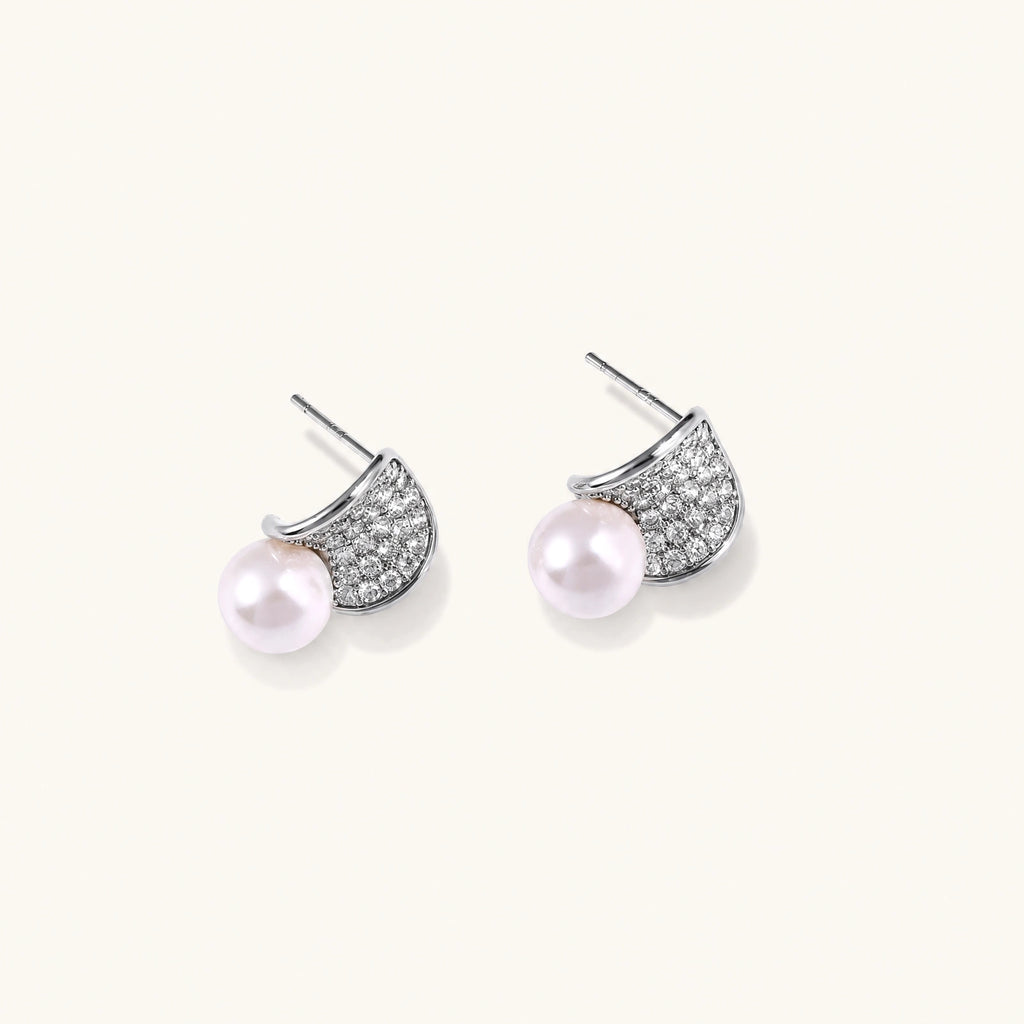Pair of white gold plated eco-brass stud earrings with curved sparkling stone panels and round freshwater pearls on a cream background.