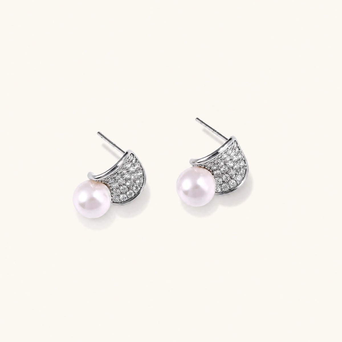 Pair of white gold plated eco-brass stud earrings with curved sparkling stone panels and round freshwater pearls on a cream background.