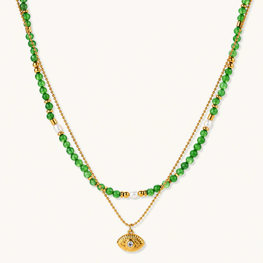 Layered necklace with green stone beads, gold spacers, white accents, and protective eye pendant on cream background.