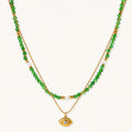 Layered necklace with green stone beads, gold spacers, white accents, and protective eye pendant on cream background.